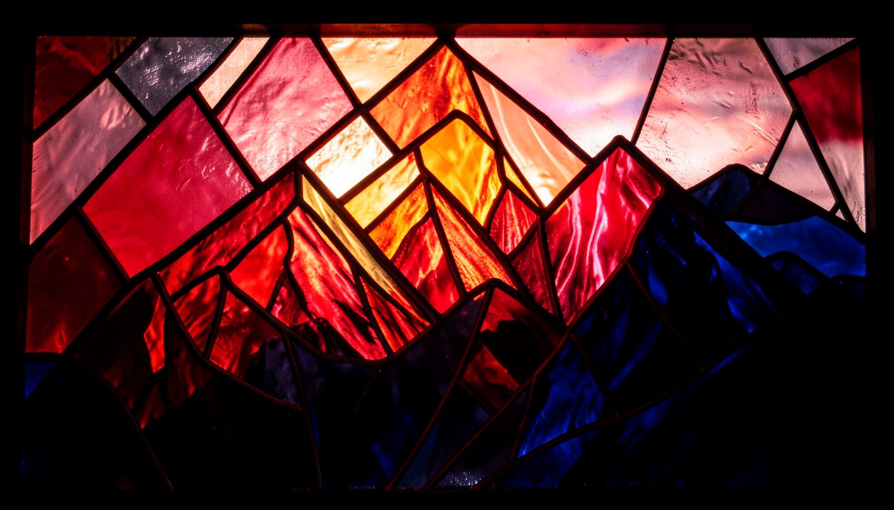 A dramatic stained-glass composition with bold leaded segments depicting mountain peaks in crimson, amber, and deep indigo